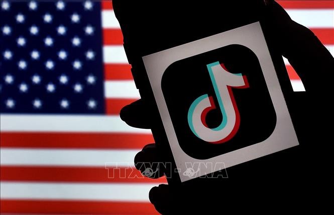 Biểu tượng ứng dụng TikTok trên màn hình điện thoại và quốc kỳ Mỹ (phía sau). Ảnh: AFP/TTXVN